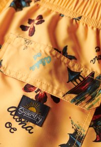 Tessuto giallo con stampe floreali e architettoniche. Presenta un'etichetta nera "Superdry" con grafica del sole e dettagli di cucitura.