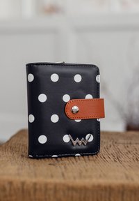 Black wallet with white polka dots, brown leather strap with snap closure, and silver accent logo on the front. Smooth texture, compact design.