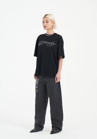 Svart oversized t-shirt med broderad "Ed Hardy"-logotyp, tillsammans med höga mörkgrå jeans prydda med färgglada fjädrar och blommönster.