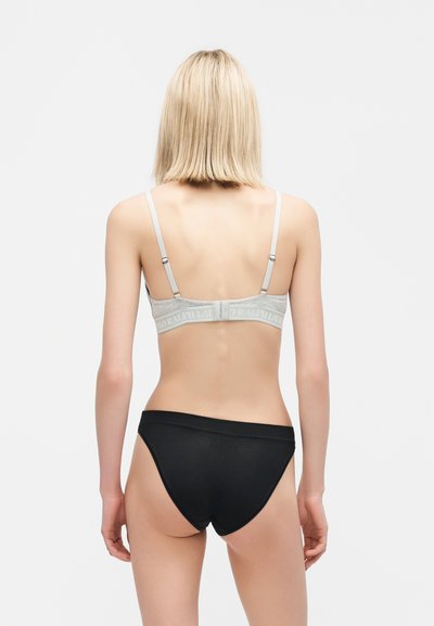 Γυναίκα με ξανθά μαλλιά που φορά ένα γκρι σουτιέν με λωρίδα λογότυπου και μαύρο εσώρουχο σε στυλ μπικίνι, εμφανίζεται από την πίσω πλευρά σε απλό φόντο.