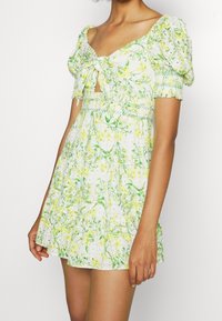 Abito floreale bianco con motivi verdi e gialli, maniche a palloncino corte, vita smock e un'apertura a chiave sul collo.