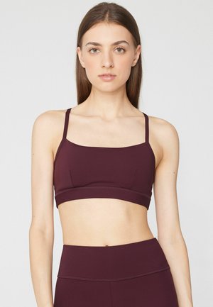 Woman with straight brown hair wearing a dark purple sports bra and matching high-waisted leggings against a plain light background.
