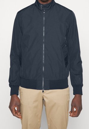 Man draagt een zwart jack met ritssluiting en drukknoopkraag en beige broek tegen een effen witte achtergrond.