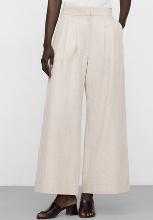 Weite beige Leinenhose, getragen mit dunkelbraunen offenen Blockabsatz-Mules, Modell steht mit einer Hand in der Tasche.