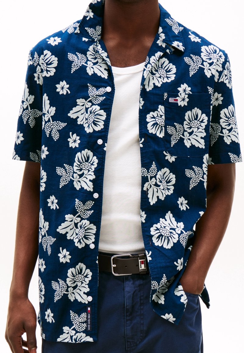 Short-sleeve button-up shirt in dark blue with white floral pattern. Features a chest pocket and a small logo tag. Made of lightweight fabric.