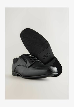 Fekete bőr öltönycipők fűzővel, sima felsőrészsel és mintázott gumitalppal, világos háttér előtt.