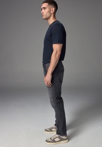 Mann steht im Profil, trägt ein marineblaues T-Shirt, dunkelgraue Jeans und grau-weiße Sneaker vor einfachem grauem Hintergrund.