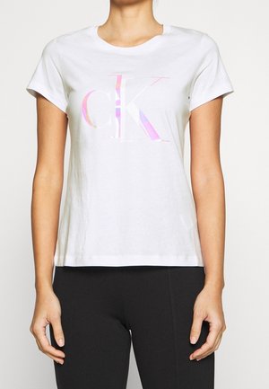 Osoba nosząca białą koszulkę z krótkim rękawem z dużym, pastelowym logo "cK" na klatce piersiowej oraz czarne spodnie.