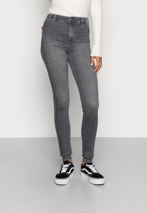 Personne portant un jean skinny gris taille haute, un top blanc à manches longues côtelées et des baskets noir et blanc, debout devant un fond uni.