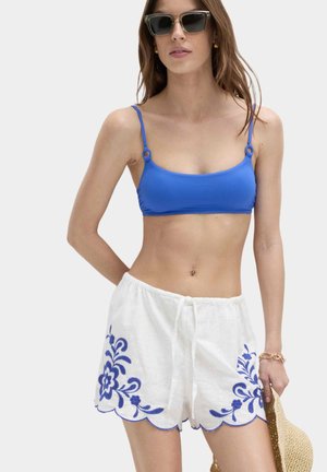 Femme portant un haut de bikini bleu, un short blanc avec une broderie florale bleue, des lunettes de soleil, et tenant un chapeau beige tissé sur un fond uni.