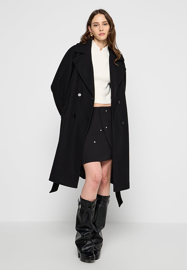 Czarny dwurzędowy trencz z szerokimi klapami, noszony na białym skrojonym swetrze i czarnej spódnicy, uzupełniony czarnymi kozakami za kolano.