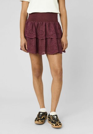 Femme portant un haut blanc, une mini-jupe à volants en dentelle bordeaux, des chaussettes blanches et des baskets à motif léopard, debout sur un fond gris uni.