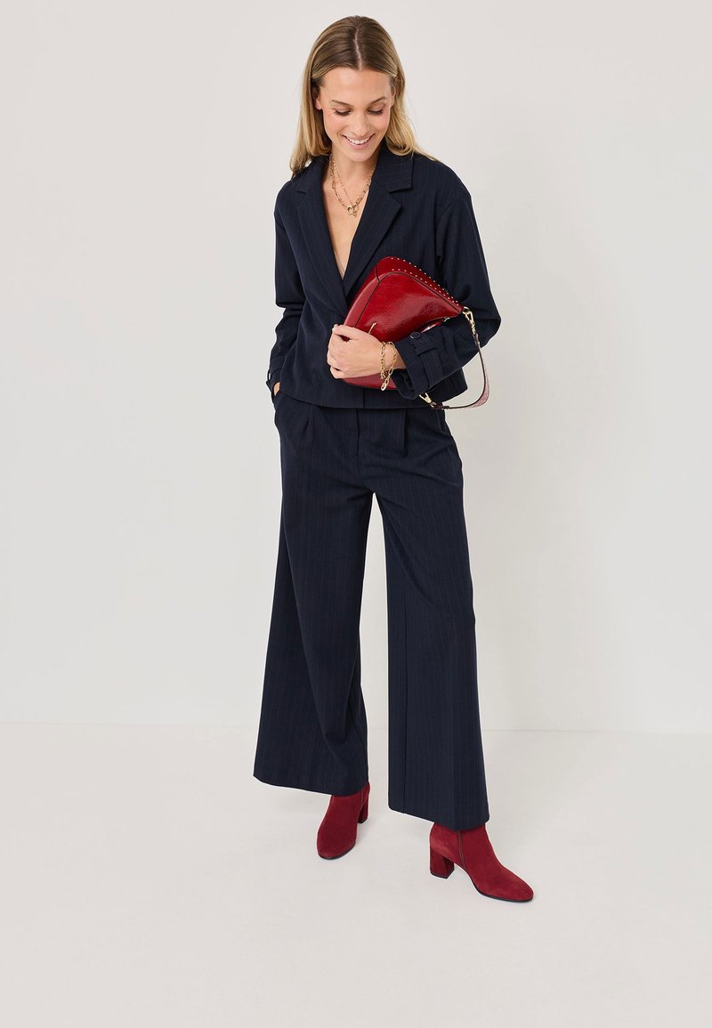 Costume rayé bleu marine avec une veste courte et un pantalon à jambes larges, associé à des bottines chevilles en daim rouge et un sac à main assorti. Accents en chaîne dorée.