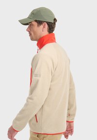 Veste en polaire crème avec des accents orange, dotée d'un col haut, de poches zippées et d'une surface texturée pour la chaleur et le confort.