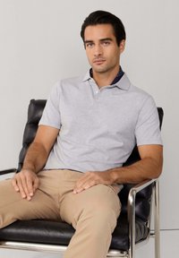 Homme aux cheveux foncés portant un polo gris clair et un pantalon beige, assis sur une chaise en cuir noire aux accoudoirs chromés, sur un fond uni.