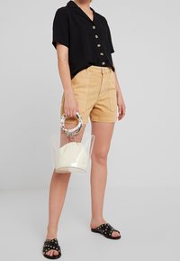 Persona che indossa una maglietta nera a maniche corte, pantaloni corti tan, sandali neri con borchie, mentre tiene una borsa trasparente con manico circolare marmorizzato e un sacchetto bianco all'interno.