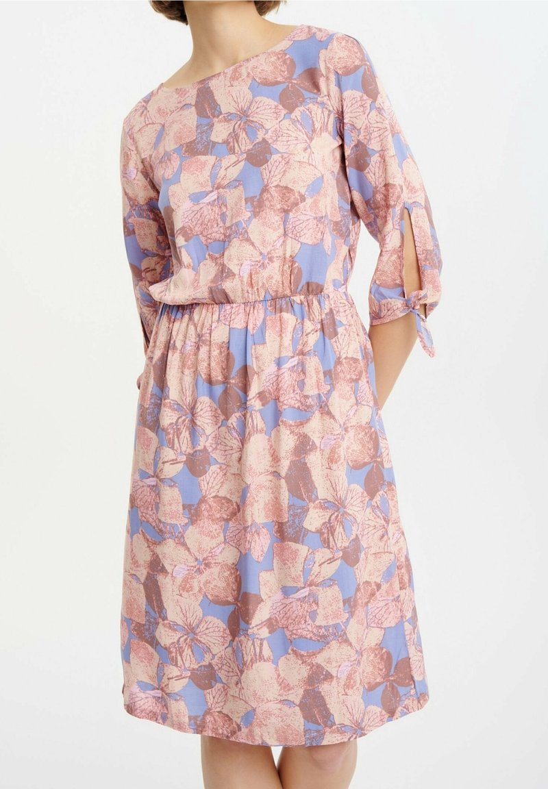 Frau in einem knielangen Kleid mit rosa- und braunfarbenen Blumenmustern auf einem helllila Hintergrund, das über Dreiviertelärmel mit gebundenen Schlitzen verfügt.