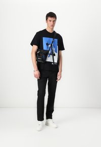 Camiseta negra con un estampado gráfico azul, combinada con pantalones negros y zapatillas blancas. Un pequeño bolso cruzado negro con detalles azules.