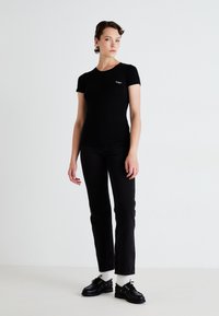 Femeie îmbrăcată într-un tricou negru ajustat, blugi negri, șosete albe și pantofi negri, așezată în fața unui fundal alb simplu.
