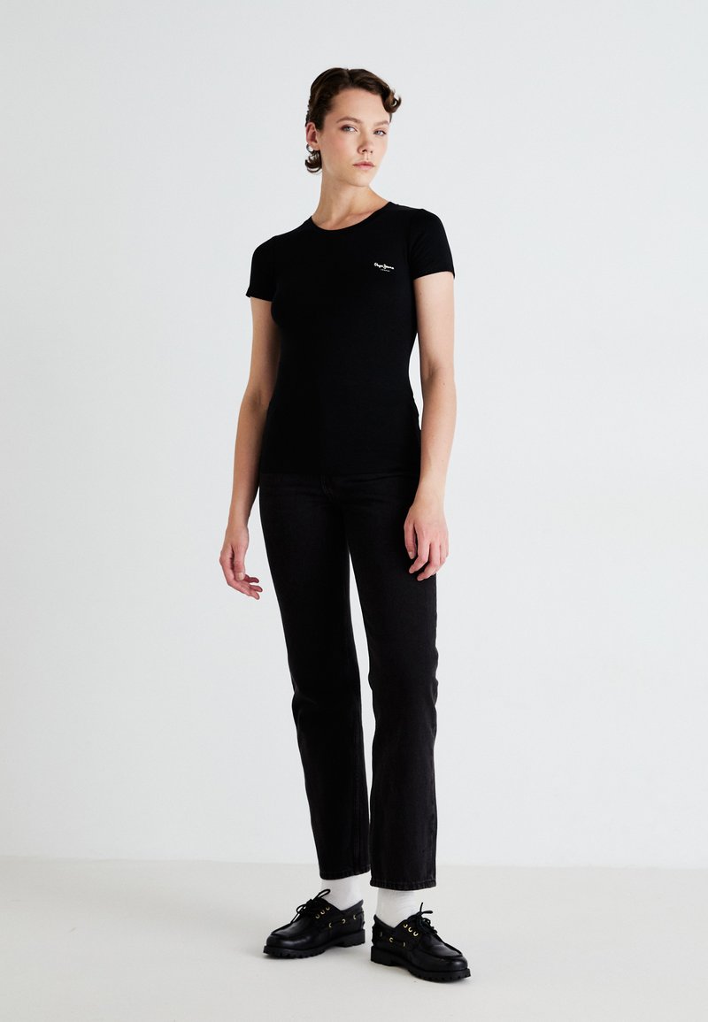 Femeie îmbrăcată într-un tricou negru ajustat, blugi negri, șosete albe și pantofi negri, așezată în fața unui fundal alb simplu.