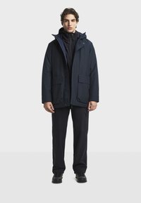 Námornícka nepremokavá bunda s kapucňou, prednými vreckami na cvoky a zipsovým zapínaním. Nosená cez tmavú základnú vrstvu a skombinovaná s tmavými nohavicami a topánkami.