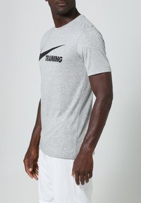 Camiseta de entrenamiento gris de Nike con mangas cortas, que presenta un logo y texto en negro en el frente, fabricada con un tejido suave y ligero.