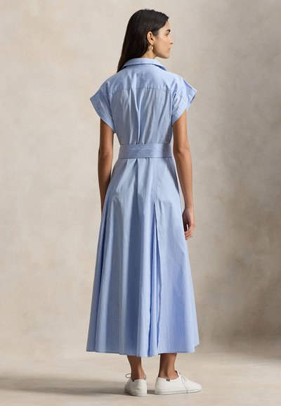 Φόρεμα ανοιχτού γαλάζιου με ρίγες, κοντά μανίκια καπέλου, ζώνη στη μέση και μακριά, αέρινη φούστα με πιέτα στο πίσω μέρος. Λευκά casual παπούτσια.