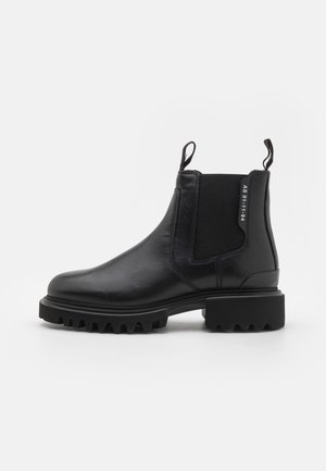 Bottine Chelsea en cuir noir avec semelle épaisse à crampons, panneaux latéraux élastiques et languettes sur le col de la cheville, texte latéral « AS 01-11-94 ».