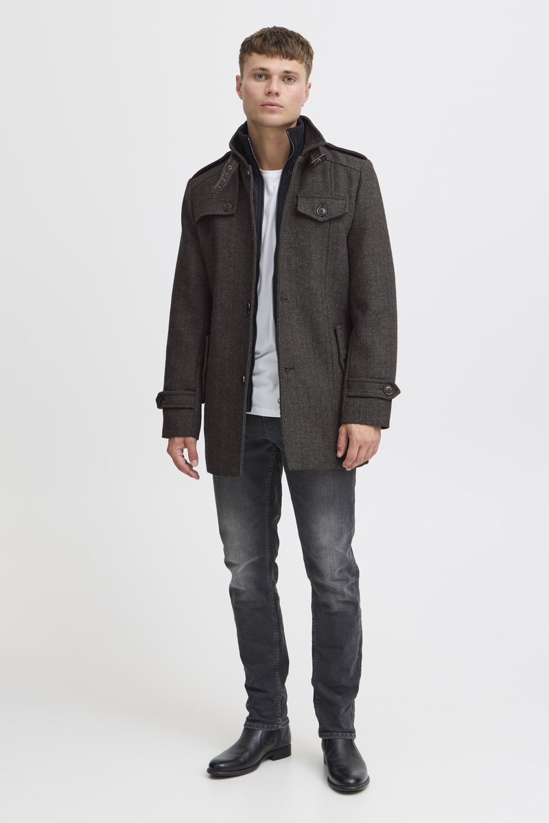 Donkergrijze wollen jas met een gestructureerd ontwerp, knopen, epauletten en zijzakken, gedragen over een wit overhemd en gecombineerd met donkere jeans en laarzen.