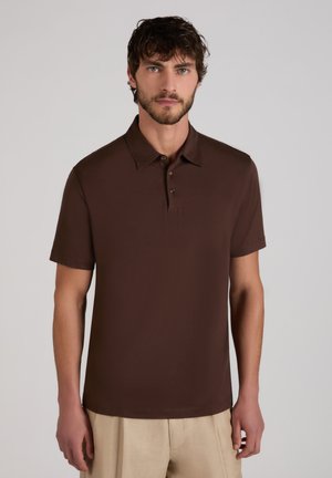 Mężczyzna o krótkich ciemnych włosach i brodzie, ubrany w brązową koszulkę polo z krótkim rękawem oraz beżowe spodnie, stojący na tle jasnego, jednolitego tła.