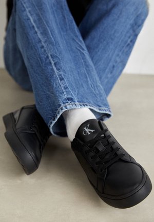 Personne assise en tailleur portant un jean bleu, des chaussettes blanches et des baskets à lacets Calvin Klein noires sur un sol neutre.