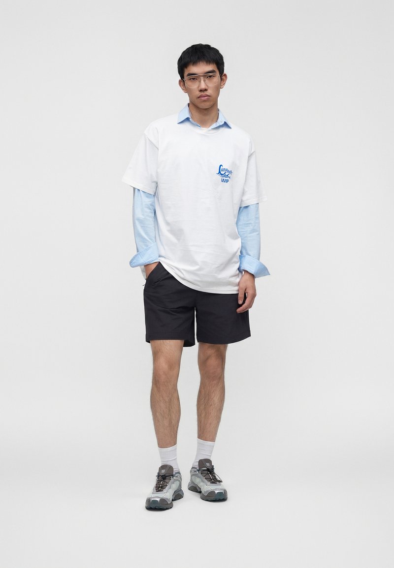 Giovane uomo con occhiali trasparenti, maglietta bianca con grafica su camicia azzurra, pantaloncini neri, calzini bianchi e sneakers grigie, in piedi con le mani parzialmente in tasca.