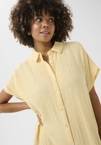 Camicia gialla con colletto e maniche corte, realizzata in tessuto testurizzato e con chiusura a bottoni. Vestibilità ampia con un drappeggio morbido e un'elegante lucentezza.