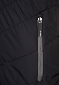 Section d'une veste matelassée noire avec une fermeture éclair grise visible. Comprend une tirette en métal et une étiquette de marque portant "Bench.". Surface texturée.