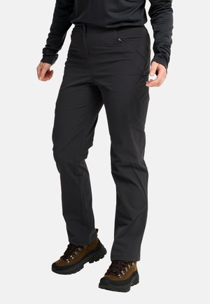 Persona che indossa pantaloni da esterno neri con tasche laterali con cerniera e scarponi da trekking marroni con suole nere e lacci rossi.
