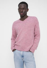 Rózsaszín V-nyakú pulóver, puha, texturált kötött anyagból, világoskék farmernadrággal párosítva. A fazon laza, bordázott mandzsettákkal és szegéllyel.