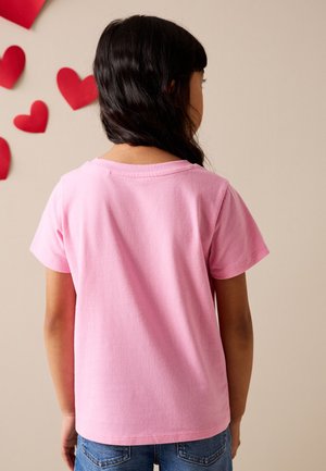 Bambino con lunghi capelli scuri che indossa una maglietta rosa semplice a maniche corte e jeans blu, di fronte a un muro beige con cuori di carta rossi.