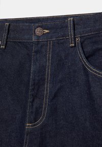Jeans en denim bleu foncé avec coutures jaunes visibles, bouton en métal, passants pour ceinture et poche avant du côté droit.