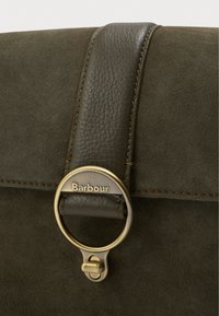 Sötétzöld velúr táska, bőrszíjjal, amely aranyszínű köralaku zárjellel van ellátva, rajta a "Barbour" felirattal. Strukturált felület és megerősített varrás.