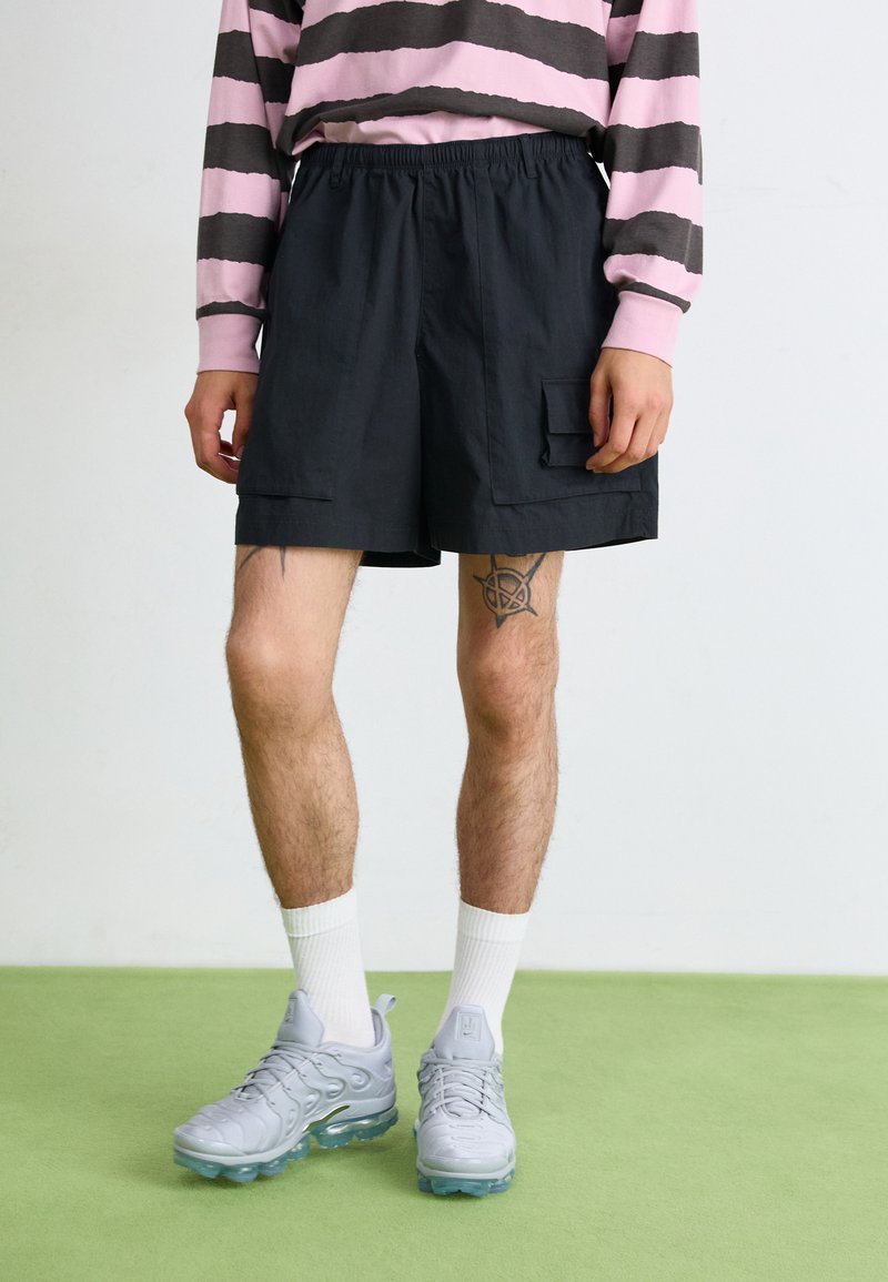 Black cargo shorts with an elastic waistband and side pockets, paired with a pink and gray striped long-sleeve shirt and gray sneakers.