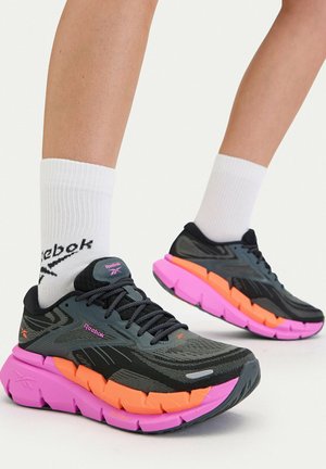 Osoba nosící černé běžecké boty Reebok s tlustými podrážkami v jasně oranžové a růžové barvě a bílé ponožky Reebok.