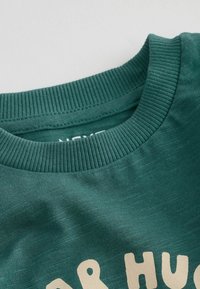 Felpa in cotone verde scuro con collo a girocollo a coste e design testuale beige. Il tessuto ha una sottile texture.