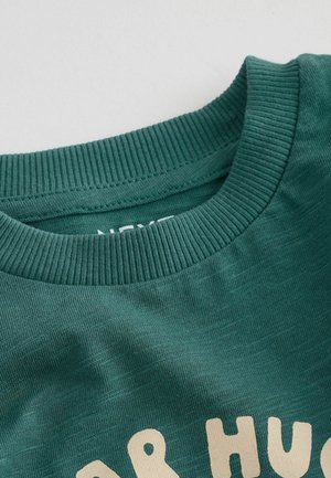Felpa in cotone verde scuro con collo a girocollo a coste e design testuale beige. Il tessuto ha una sottile texture.