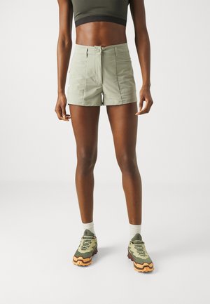 Frau trägt hellgrüne Shorts mit hohem Bund, dunkelgrünes kurzes Top, weiße Socken und grün-orange Wanderschuhe, steht vor weißem Hintergrund.