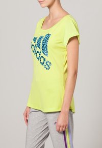 Ljusgul kortärmad t-shirt med en blårandig prickig Adidas-logotyp över bröstet. Mjukt tyg. Bärs med gråa mjukisbyxor med lila ränder.