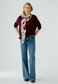 Weinroter Strickpullover mit tiefem V-Ausschnitt, kombiniert mit weit geschnittenen blauen Jeans. Ein leichter Schal mit einem auffälligen Muster in Rot und Beige.