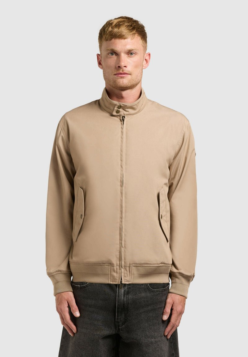 Homme portant une veste beige à fermeture éclair avec col haut et poches latérales à rabat, associée à un jean gris foncé, debout devant un fond uni.