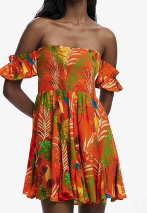 Kobieta ubrana w pomarańczową sukienkę odkrywającą ramiona z zielonymi, żółtymi, niebieskimi i białymi tropikalnymi liśćmi i kwiatowymi wzorami, stojąca z rękami za plecami.