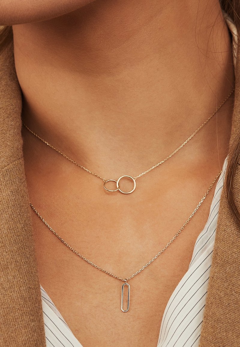 Deux colliers délicats en chaîne dorée, l'un avec deux anneaux entrelacés et l'autre avec un pendentif ovale vertical, portés au cou d'une femme.