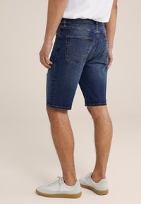 Tamsiai mėlynos džinsinės šortai, siekiantys vidurinę šlaunų dalį, su užpakalinėmis kišenėmis, kurių siūlės matomos. Dėvimi su šviesiai pilkais sportbačiais.